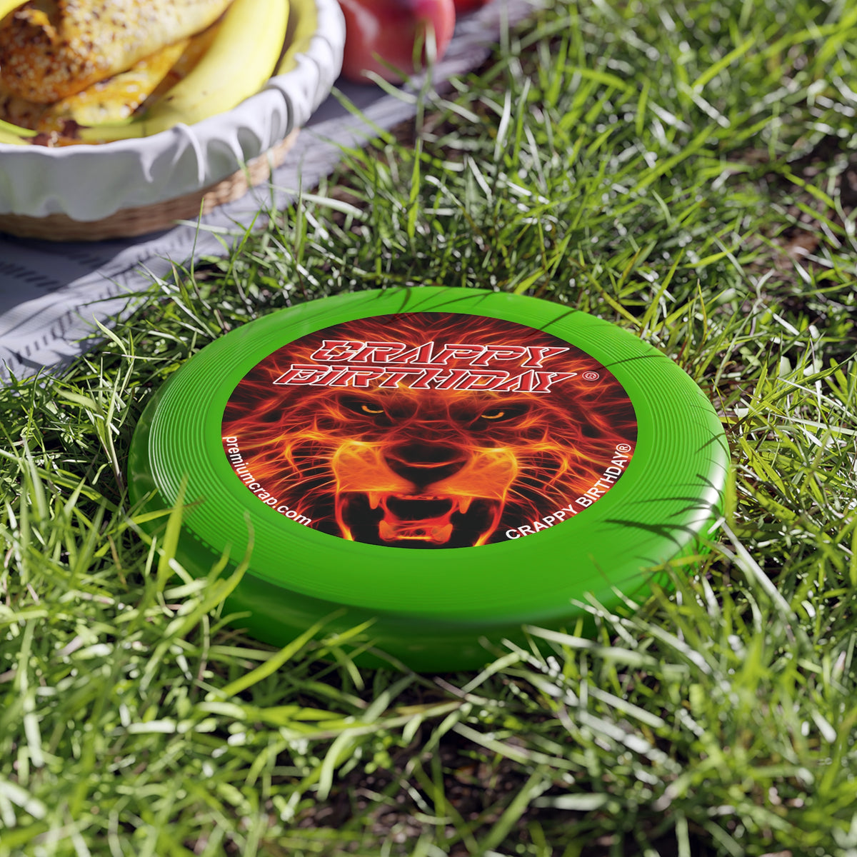 A green plastic flying disc with a red and orange fiery design and text 'CRAPPY BIRTHDAY' laid on grass.