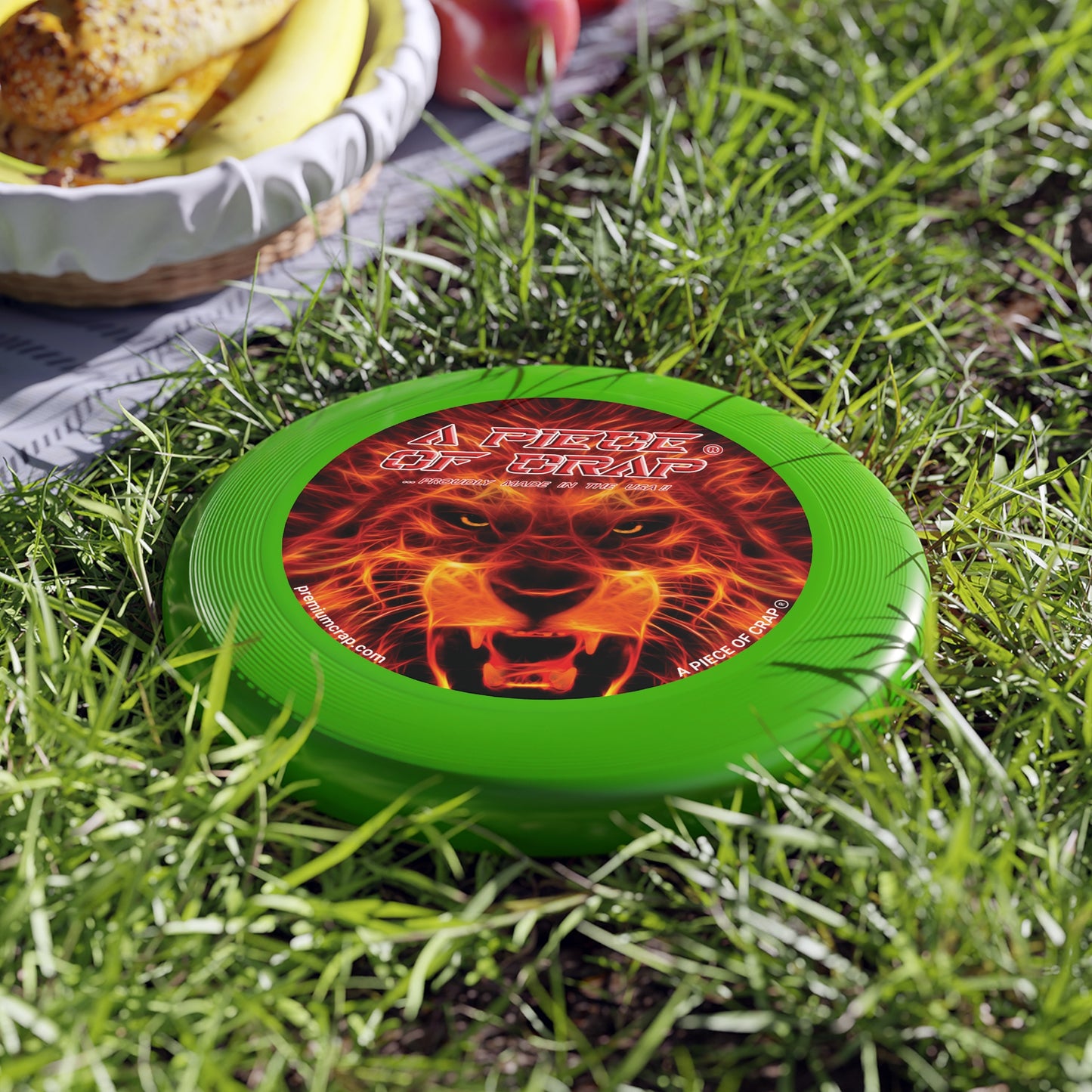 A green flying disc with a red and orange flame-like design and text that reads 'A Piece of Crap' lying on grass next to a burger and fries.