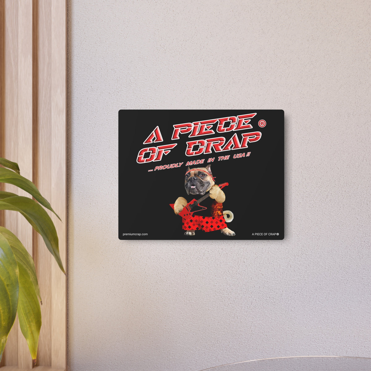 A square black wall art print featuring a pug in a red polka-dot dress with the text 'A PIECE OF CRAP' in red and white, mounted on a light gray wall next to a plant.