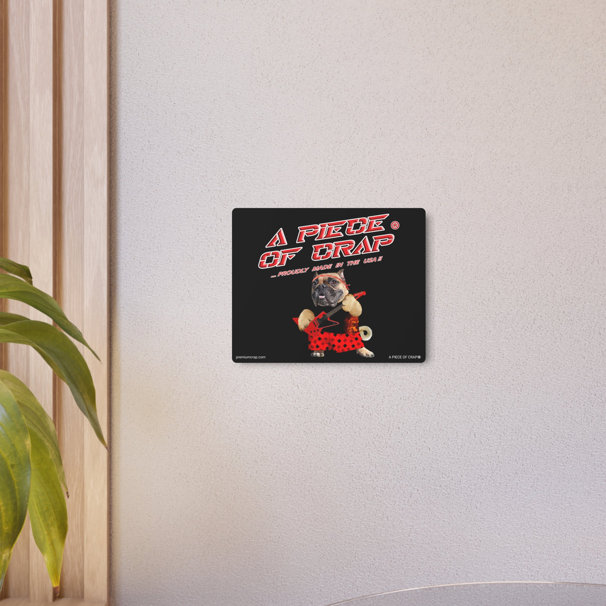 A rectangular black wall art print mounted on a light gray wall, featuring the text 'A PIECE OF GRAP' in red and white above a cartoon pug dressed in a red polka-dot outfit. A plant and wooden trim are visible at the left edge of the scene.