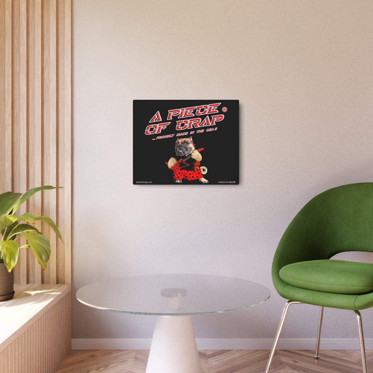 A square wall art print featuring a retro-style graphic with the text 'A PIECE OF CRAP' and a cartoon dog in a red polka-dot outfit, displayed on a light beige wall in a modern room setting with a green chair, glass table, and wood slat paneling.