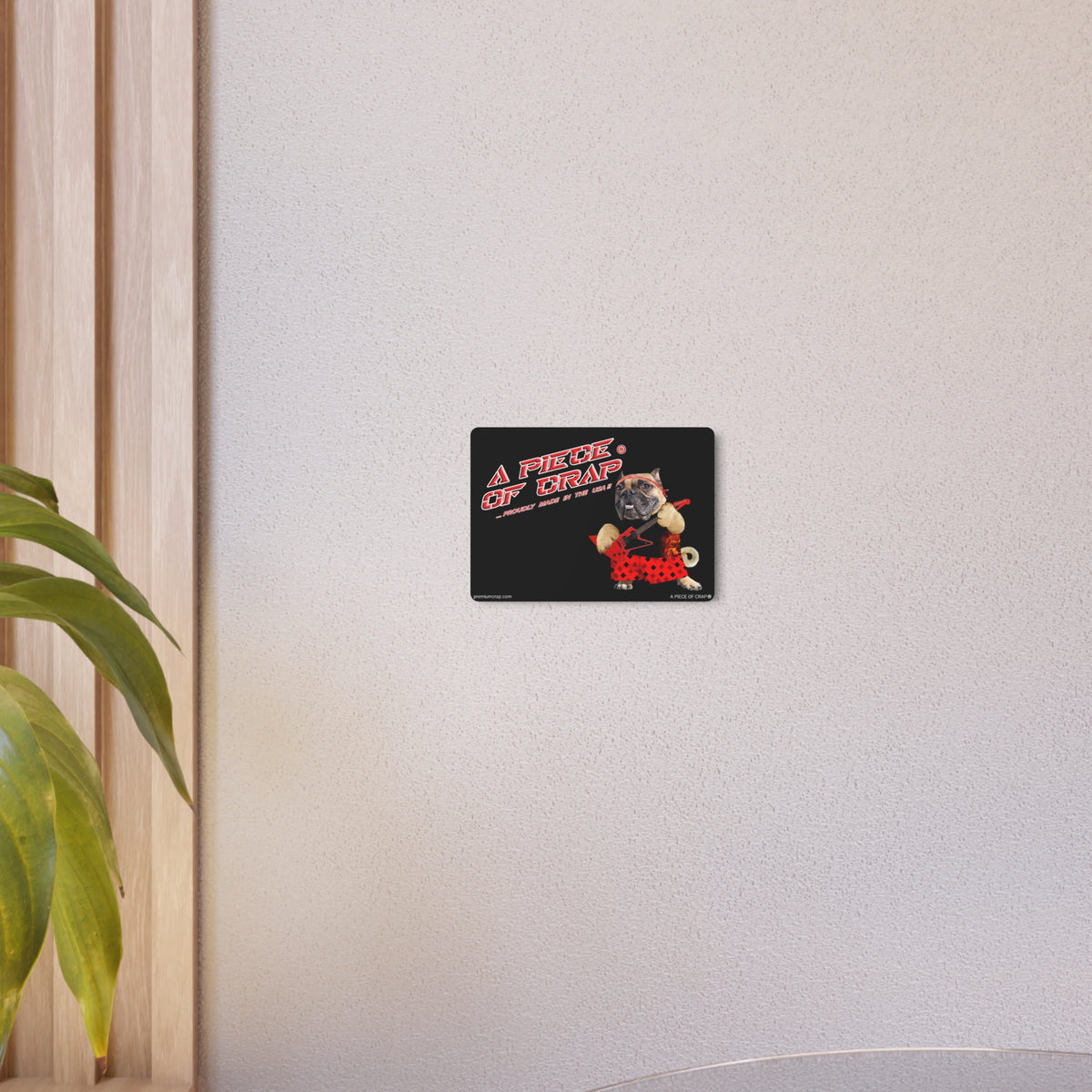 A rectangular black wall sticker or decal with the word A Piece of Crap' in white at the top, shown mounted on a light beige textured wall. A green plant leaf is visible on the left side of the frame.