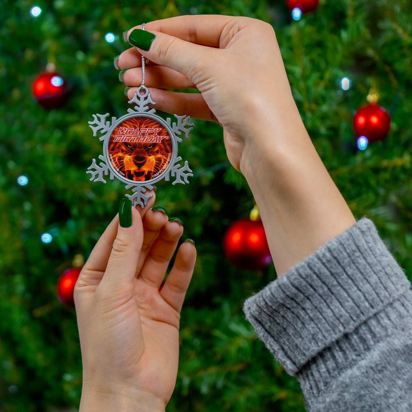A person holds a silver snowflake Christmas ornament with a red center featuring a lion and the text 'Crappy Birthday' in front of a blurred Christmas tree with red ornaments.