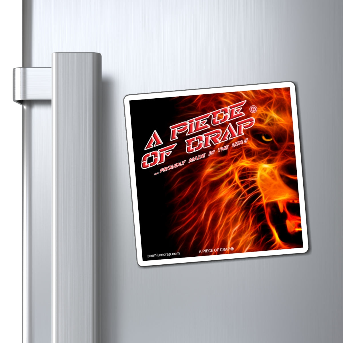 A rectangular refrigerator magnet with a white border, featuring a fiery lion graphic and the text 'A PIECE OF CRAP - PROUDLY MADE IN THE USA', shown attached to a brushed stainless steel refrigerator door next to the handle.