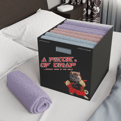 A black storage box labeled 'A PIECE OF CRAP' with a dog graphic, filled with folded towels in pastel colors, shown on a bed next to a rolled lavender towel.